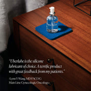 A clear bottle of UberLube Premium Silicone Lubricant 50 ml with a pump rests on a blue coaster on a wooden dresser, with an endorsement by Dr. Lynn Y Wang shown in the lower left corner.