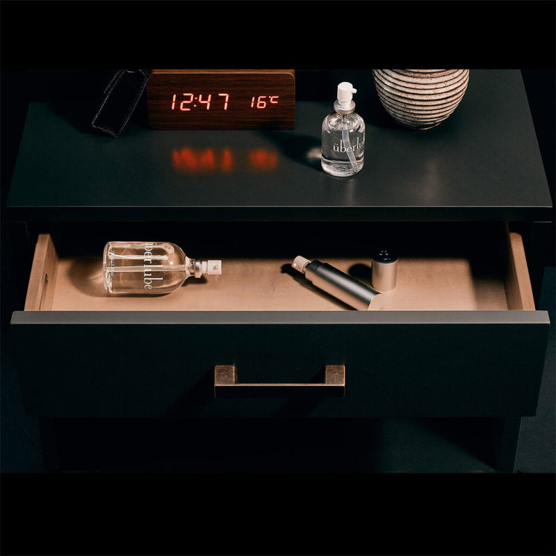 A bedside table with an open drawer. On top are a digital clock (12:47, 16°C), a small bottle, and a striped vase. Inside the drawer are UberLube Premium Silicone Lubricant 50 ml and a cylindrical object, possibly a vibrator.