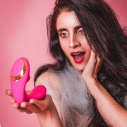 A woman with long dark hair and red lipstick looks surprised and intrigued as she holds Tracy's Dog OG Clitoral Sucking G-Spot Vibrator against a pink background.