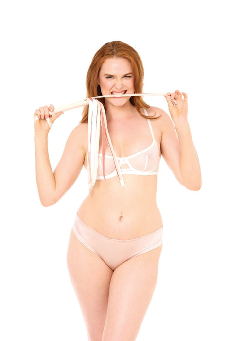 A woman in pale lingerie playfully bites a strap while holding the Peaches 'n Cream Flogger in both hands, standing against a plain white background.