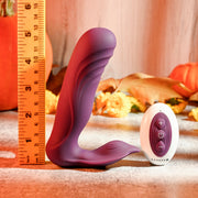 The Velvet Hammer Thrusting Hands-Free Dual Stimulation Vibrator, a purple L-shaped waterproof silicone toy with textured curves, is displayed next to a ruler and white remote, on a surface decorated for fall.