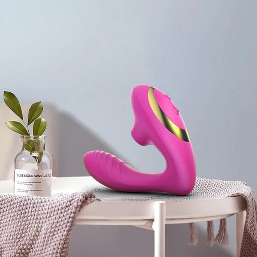 Tracy's Dog OG Clitoral Sucking G-Spot Vibrator in pink and gold rests on a white round table next to a glass vase with green leaves and a gray textured cloth, set against a light blue wall.