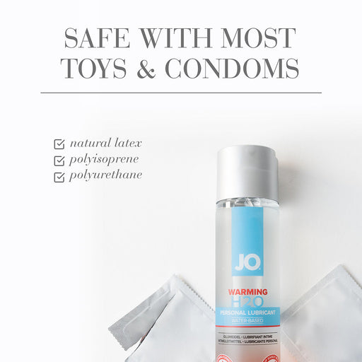 A bottle of Jo H2O Warming Water Based Lubricant stands upright beside two unwrapped condoms on a white surface. Above, text reads: "SAFE WITH MOST TOYS & CONDOMS" and checks off natural latex, polyisoprene, and polyurethane.