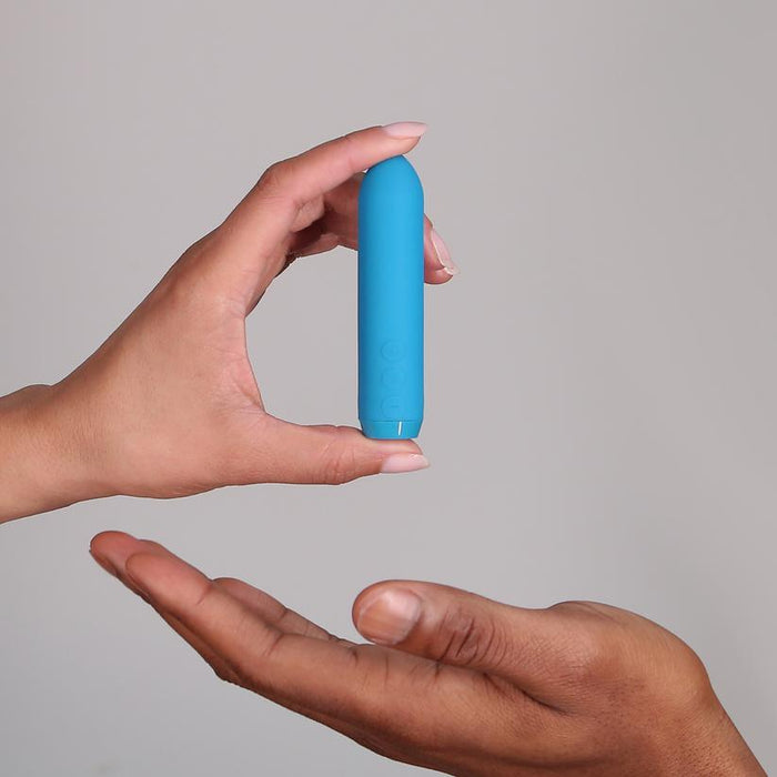 Two hands are shown; one holds the Je Joue Powerful Classic Bullet with Finger Sleeve in teal, while the other is open beneath it against a plain light background.