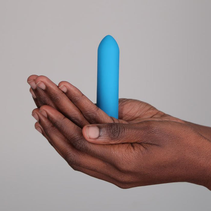 A person with dark skin holds the Je Joue Powerful Classic Bullet with Finger Sleeve in teal between both hands, showing its small, USB rechargeable design and rumbly vibrations against a neutral background.