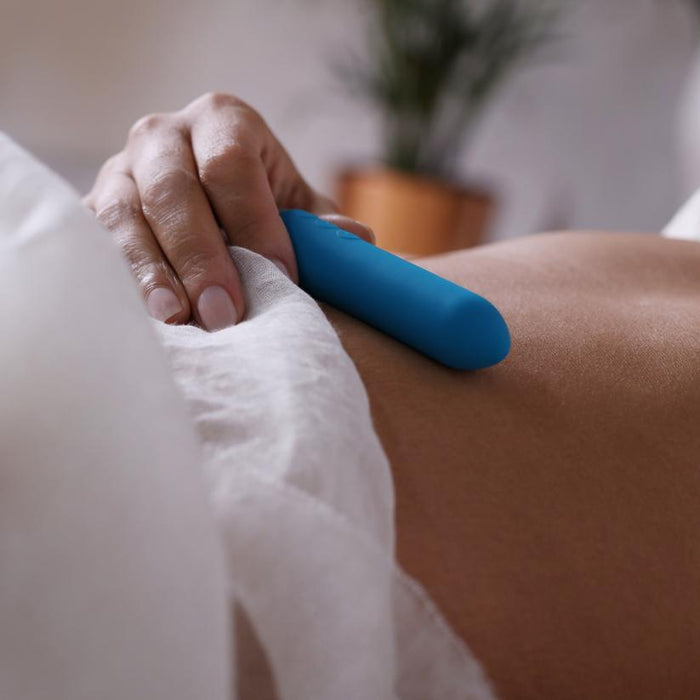 A person lies down, a hand holding the Je Joue Powerful Classic Bullet with Finger Sleeve - Teal against their bare abdomen under a white sheet. A blurred plant adds softness to the background.