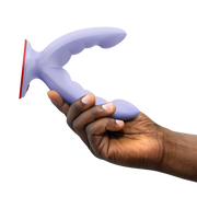 Hand holding a lavender silicone grinding dildo with a curved, ridged design and red suction base.