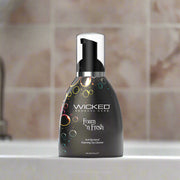 A black bottle of Wicked Foam N Fresh Toy Cleaner 8oz sits on a bathroom counter with a beige tiled wall in the background. The alcohol-free, anti-bacterial cleaner features colorful bubble graphics on the label.