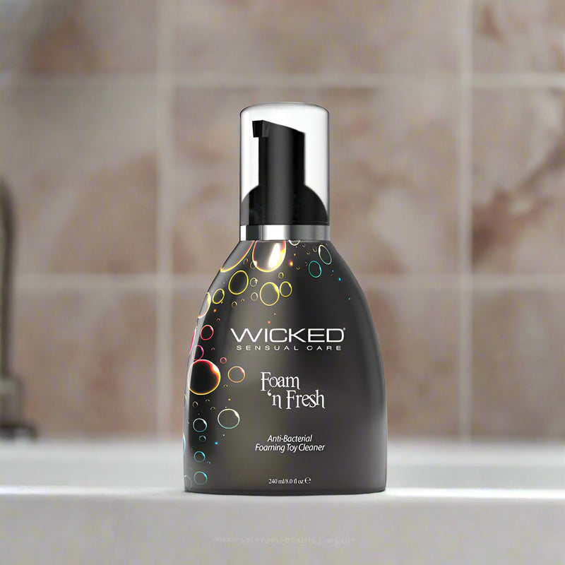 A black bottle of Wicked Foam N Fresh Toy Cleaner 8oz sits on a bathroom counter with a beige tiled wall in the background. The alcohol-free, anti-bacterial cleaner features colorful bubble graphics on the label.