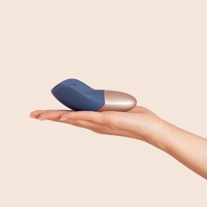 A hand with light skin tone holds The Arouser Pulsating External Stimulator in blue and metallic colors, made from body-safe silicone, against a plain beige background.