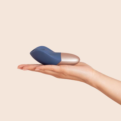 A hand with light skin tone holds The Arouser Pulsating External Stimulator in blue and metallic colors, made from body-safe silicone, against a plain beige background.