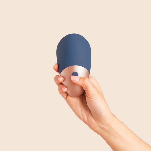 A hand holds The Arouser Pulsating External Stimulator, a compact oval blue and metallic device with a single button, made from body-safe silicone, against a plain beige background.