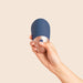 A hand holds The Arouser Pulsating External Stimulator, a compact oval blue and metallic device with a single button, made from body-safe silicone, against a plain beige background.