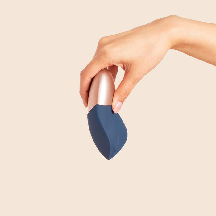 A hand holds The Arouser Pulsating External Stimulator, a modern device with body-safe silicone and blue-silver finishes, against a beige background.