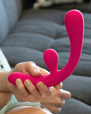 A close-up of a person's hands holding a Jimmyjane Reflexx Rabbit 3 First Time Slim Flexible Warming Vibrator - Pink by Pipedream Products with a smaller protrusion, possibly a rabbit vibrator for g-spot stimulation, in a living room setting.