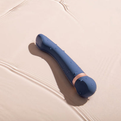 The Hot & Cold Temperature-Changing G-Spot Massager, featuring blue silicone and a gold accent, rests on a smooth beige surface with water droplets—ideal for temperature play and relaxation.