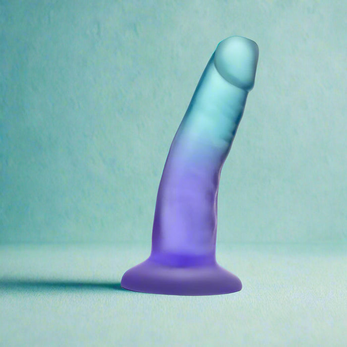 The Morning Dew Small 5 Inch Dildo with Suction Cup - Blue, a translucent gradient blue and purple dildo with a flared base, stands upright on a light teal surface. It's beginner-friendly, suction-based, and harness compatible for versatile play.