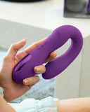 A close-up image of a hand holding a purple, u-shaped silicone device, likely a Jimmyjane Reflexx Rabbit 1 G-Spot & Clit Hugging Warming Vibrator - Purple with dual independently controlled motors. The background is softly blurred, focusing attention on the object.
