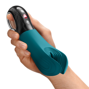 Hand holding a teal silicone Fun Factory Manta penis stroker with a black textured handle and red control buttons.