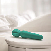 The Petite Private Pleasure Waterproof Silicone Wand Vibrator, in teal silicone, rests on a white round bedside table with a softly blurred bed and white bedding in the background.