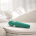 The Petite Private Pleasure Waterproof Silicone Wand Vibrator, in teal silicone, rests on a white round bedside table with a softly blurred bed and white bedding in the background.