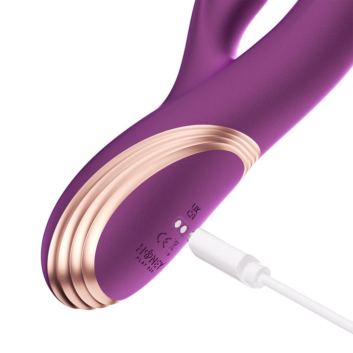 A close-up of the Gem Pressure Sensing App-Controlled Twin Balls Rabbit (Damaged Box), a purple and gold silicone massager with pressure sensing technology, being charged via a white USB cable. Charging port and indicator lights are visible.