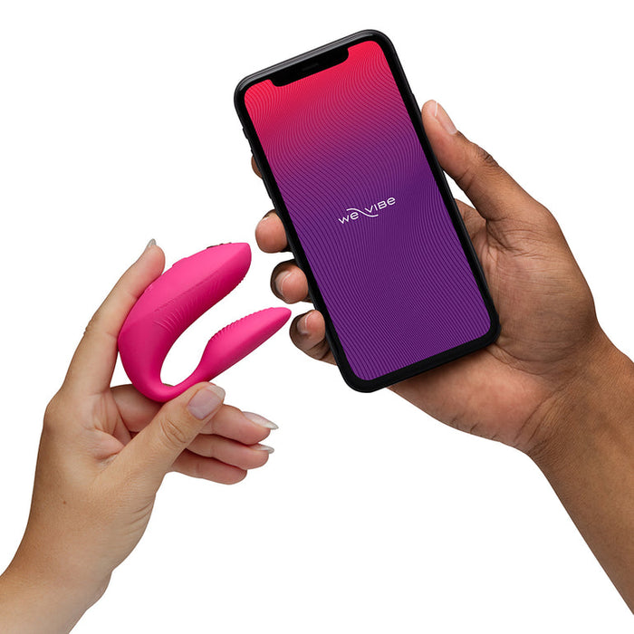 A person holds the We-Vibe Chorus Pro Wearable Couples' Vibrator with Triple Motors & Remote, while another hand shows a smartphone displaying the We-Vibe app.