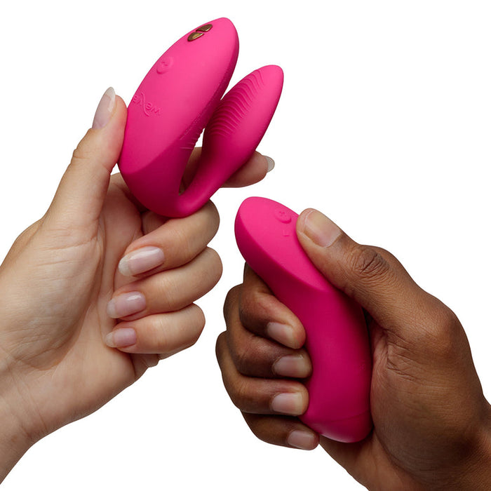 Light- and dark-skinned hands hold the We-Vibe Chorus Pro Wearable Couples' Vibrator with Triple Motors & Remote—one C-shaped for G-spot stimulation, the other elongated—both feature Fusion Wave Technology, shown on a white background.