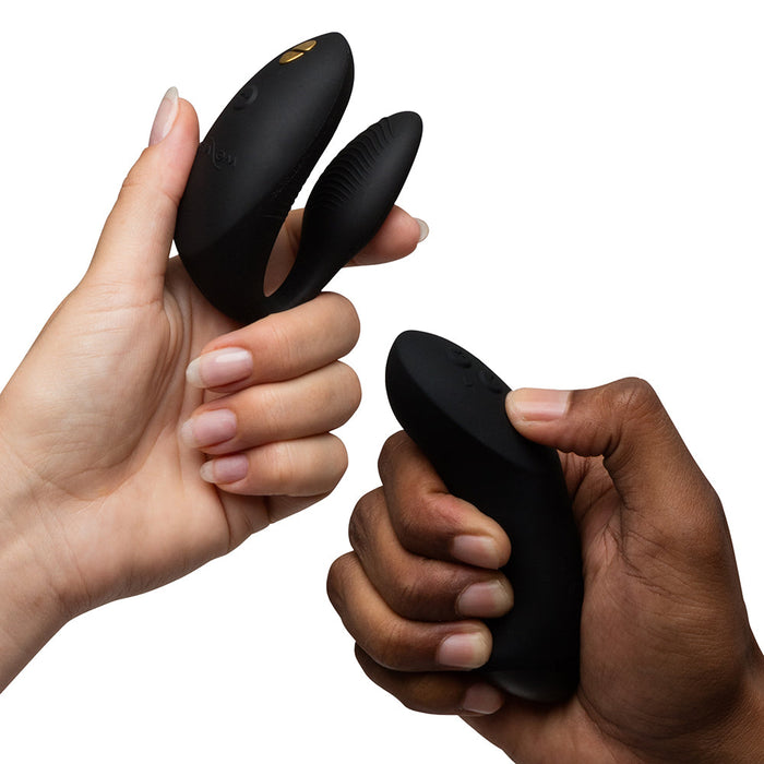 Two hands hold the black We-Vibe Chorus Pro Wearable Couples' Vibrator with triple motors and its buttoned remote against a white background.