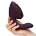 A hand holds the Womanizer Next Duo Pleasure Air + Vibration Blended Orgasm Stimulator, a dark purple, curved device with a rounded end and ergonomic shape, set against a white background.