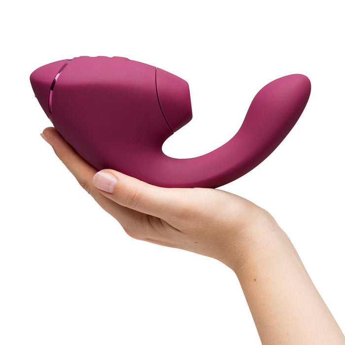 A hand holds the Womanizer Next Duo Pleasure Air + Vibration Blended Orgasm Stimulator, a burgundy, ergonomically designed G-spot vibrator with a smooth texture, photographed against a plain white background.