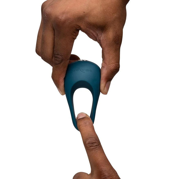 A hand holds the teal We-Vibe Pivot 2 Vibrating Silicone Rechargeable Penis Ring, designed for hands-free clitoral stimulation, above an upward-pointing finger against a white background. "We-Vibe" is visible on the silicone ring.