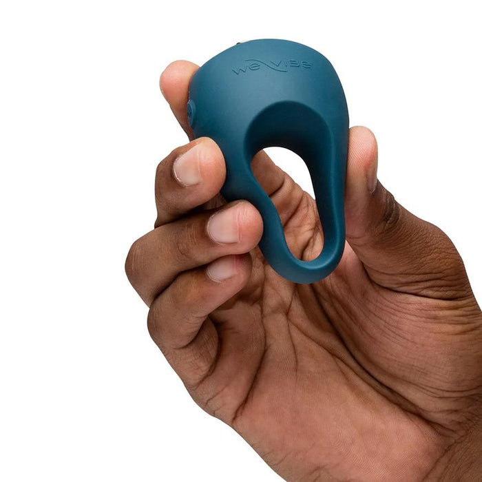 A hand holds the blue We-Vibe Pivot 2 Vibrating Silicone Rechargeable Penis Ring, with "we-vibe" branding visible against a white background.
