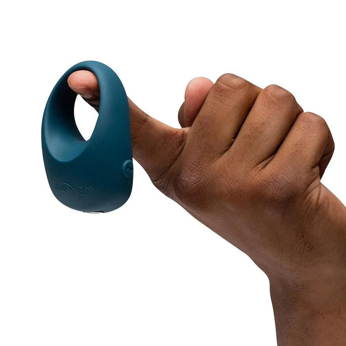 A hand with medium brown skin holds up the teal We-Vibe Pivot 2 Vibrating Silicone Rechargeable Penis Ring, designed for hands-free clitoral stimulation, isolated on a white background.