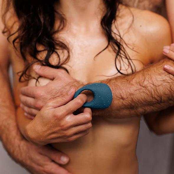 A shirtless couple embraces; one presses the We-Vibe Pivot 2 Vibrating Silicone Rechargeable Penis Ring—a blue, waterproof pleasure device—against the other’s forearm, hinting at hands-free clitoral stimulation and an intimate, sensual moment.