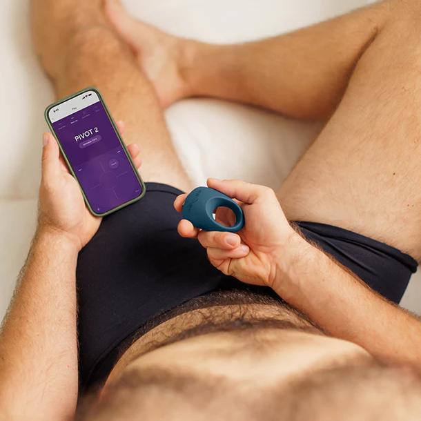 A person in black underwear lies on a bed, holding a smartphone with an app open and the We-Vibe Pivot 2 Vibrating Silicone Rechargeable Penis Ring in their other hand.