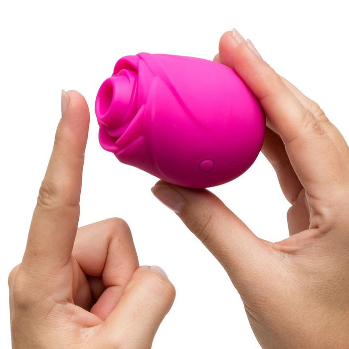 Someone holds the Romp Rose 2 Pleasure Air Clitoris Stimulator in pink—a rose-shaped, rechargeable silicone sex toy—in one hand and points at it with the other, set against a white background.