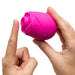 Someone holds the Romp Rose 2 Pleasure Air Clitoris Stimulator in pink—a rose-shaped, rechargeable silicone sex toy—in one hand and points at it with the other, set against a white background.