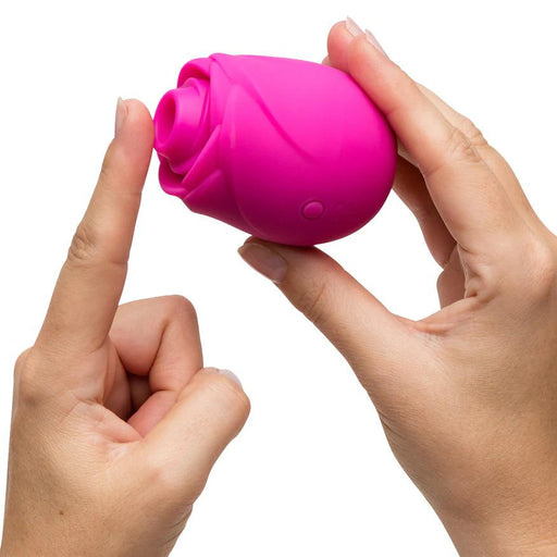 Two hands hold the Romp Rose 2 Pleasure Air Clitoris Stimulator in pink—a rechargeable, rose-shaped silicone toy with a small button—commonly used for clitoral stimulation, shown against a white background.