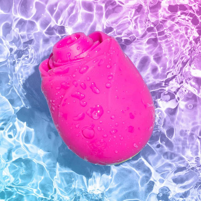 The Romp Rose 2 Pleasure Air Clitoris Stimulator in pink, shaped like a rose with water droplets, floats in clear blue and purple water reflecting shimmering light.