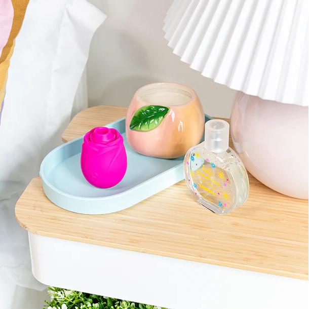 A bedside table with a pastel striped pillow, a blue tray holding the Romp Rose 2 Pleasure Air Clitoris Stimulator in pink, a candle with a green leaf, a heart-shaped bottle with gold stars, and a white lamp.