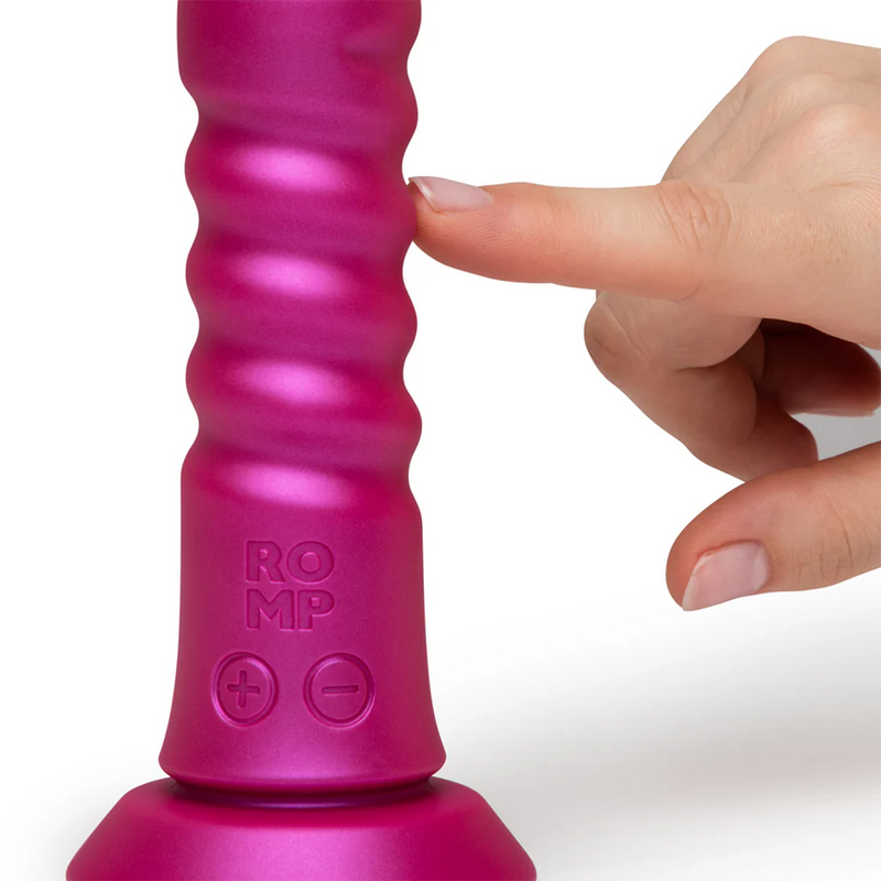 Pink ROMP Jiggle thrusting vibrator with a ribbed silicone shaft, suction base, and control buttons, being touched by a finger.