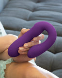 A person holding a Pipedream Products Jimmyjane Reflexx Rabbit 1 G-Spot & Clit Hugging Warming Vibrator in purple, sitting on a grey couch. The focus is on the device and hand with a glimpse of light-colored clothing.
