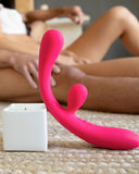 A Jimmyjane Reflexx Rabbit 3 First Time Slim Flexible Warming Vibrator in pink on a textured surface, with a blurred background of a person seated on a bed. A white candle is in the foreground.