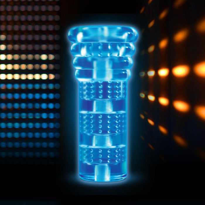 A glowing blue, cylindrical object with multiple stacked segments and textured surfaces, resembling Blush's All Nighter Glow in the Dark Textured Clear Self Lubricating Stroker. It stands against a dark background illuminated by rows of orange and blue lights, creating a high-tech ambiance.