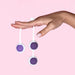 A hand with light pink nail polish balances two white and purple balls from the Wellness Kegel Ball Training Kit for Pelvic Floor Health on its fingers, set against a solid light pink background.