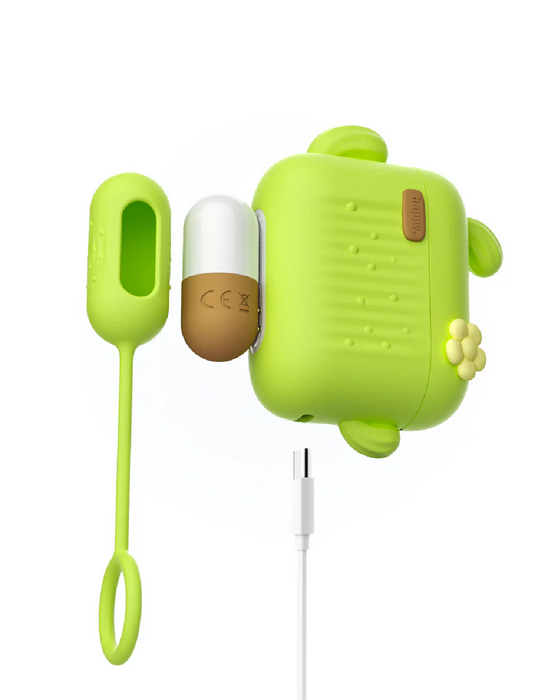 A green, turtle-shaped electronic device with a small brown cap on its side is connected to a charging cable. A matching green silicone loop attachment is placed near the device. This Blooming Bliss First Time Bullet & Egg Vibrators with Charging Case & Remote by Blush features button-like details and a small flower decoration on the side.