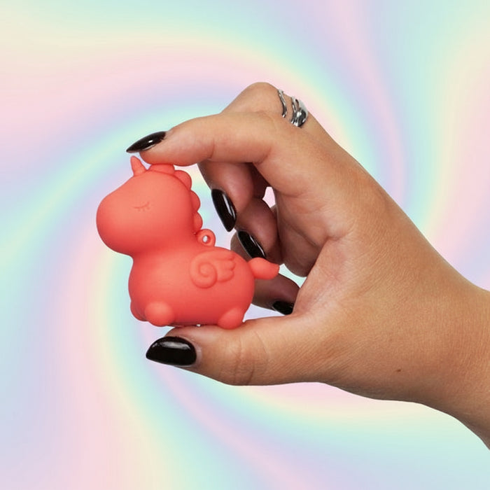 A hand with black-painted nails holds the Unihorn Mini - Peachy Pony Discreet Vibrator, a small pink silicone unicorn, against a pastel rainbow swirl background.