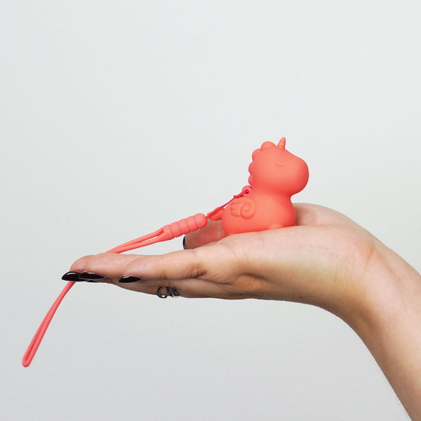 A hand with black nail polish holds the Unihorn Mini - Peachy Pony Discreet Vibrator, a small, unicorn-shaped toy made of body-safe silicone with a wrist strap, set against a plain white background.
