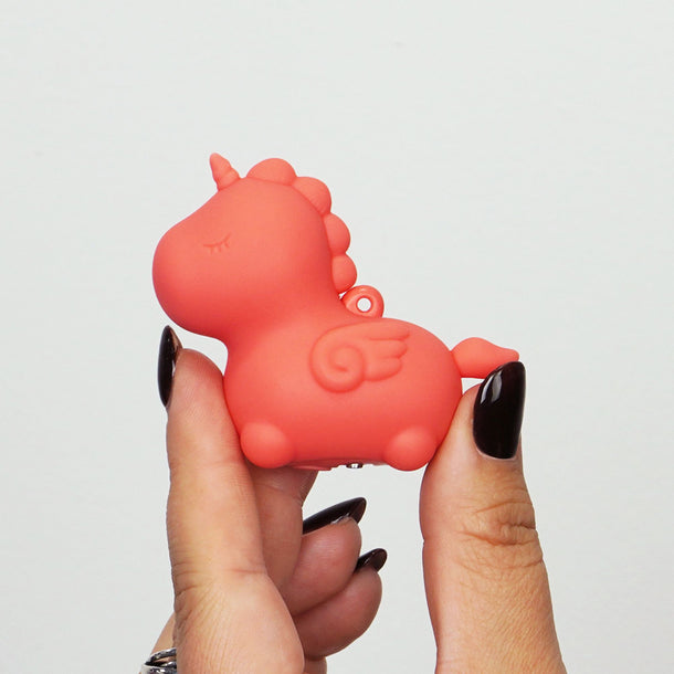 A hand with black-painted nails holds the Unihorn Mini - Peachy Pony Discreet Vibrator, a small red unicorn-shaped vibe with wings and a closed-eye design, made of body-safe silicone, against a plain white background.